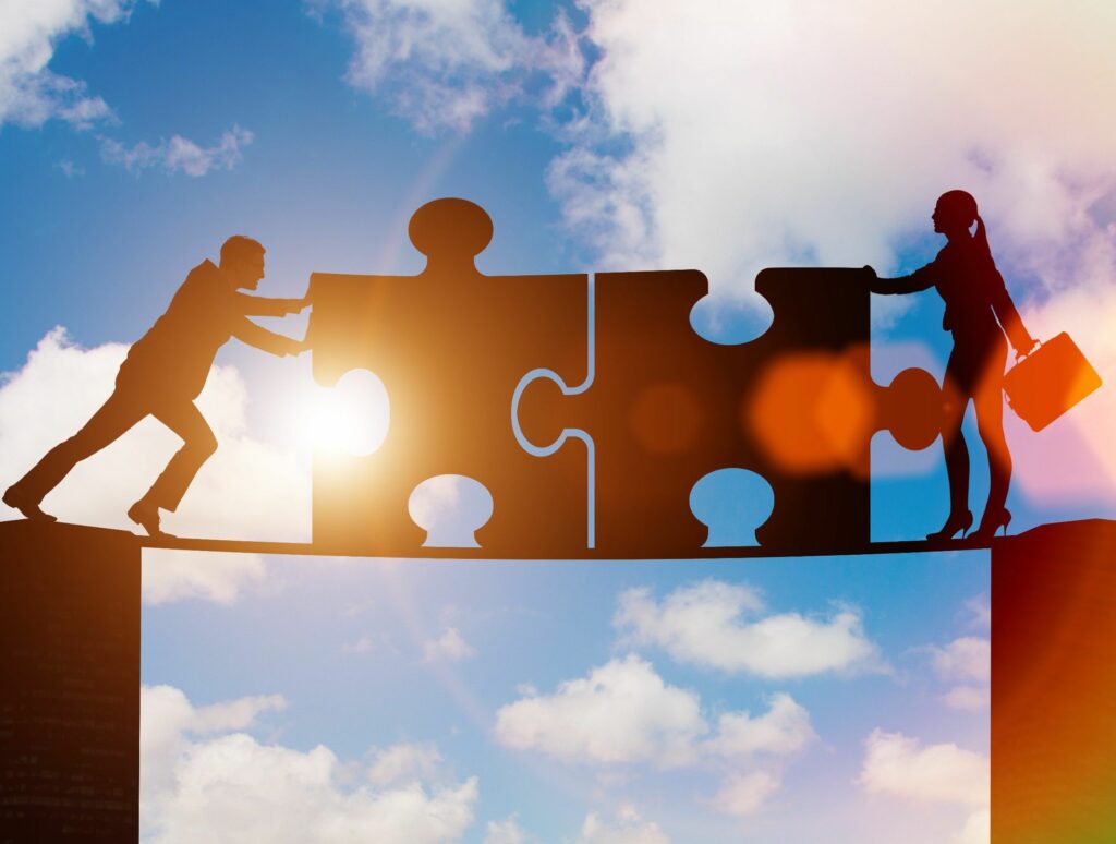 Silhouettes of a man and woman pushing large puzzle pieces together against a bright sky, symbolizing collaboration and problem-solving.