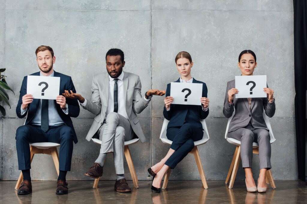 Four professionals in suits sit on chairs, each holding a sign with a question mark, expressing confusion or uncertainty.