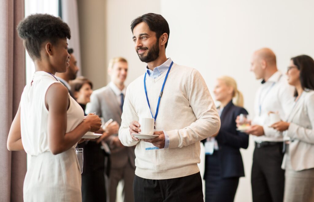 Two professionals engage in conversation at a networking event, surrounded by others, all holding drinks and plates, creating a collaborative atmosphere.
